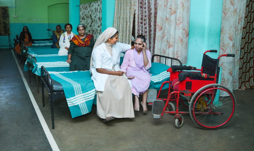 A religious sister sits next to and embraces a woman in a hospital. There is a wheelchair next to the bed.