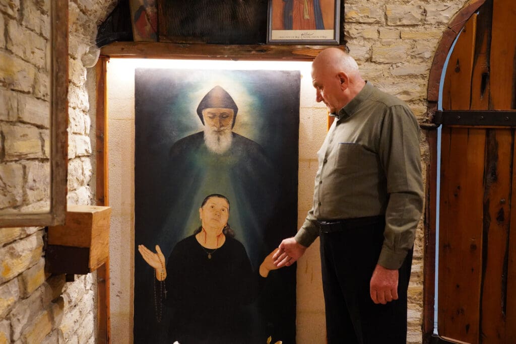 A man stands by a painting depicting St. Charbel and a woman.