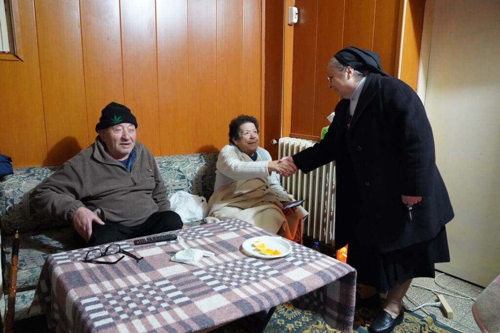 A religious sister shakes the hand of a woman sitting on a couch. Another man is sitting next to the woman.