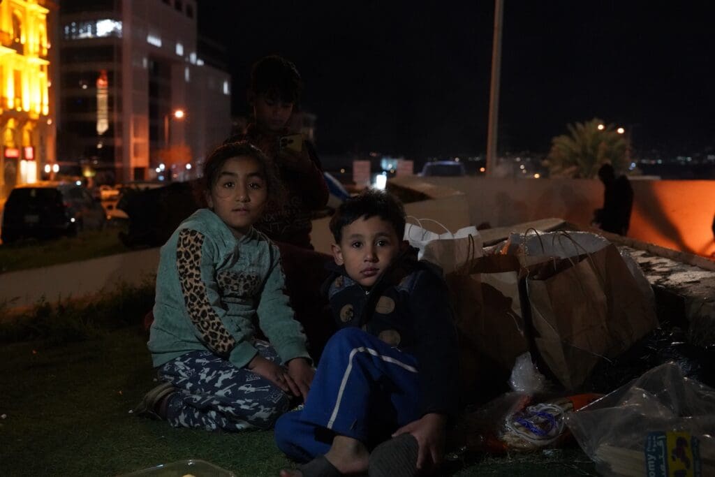 Two children sitting on a street next to cardboards.
