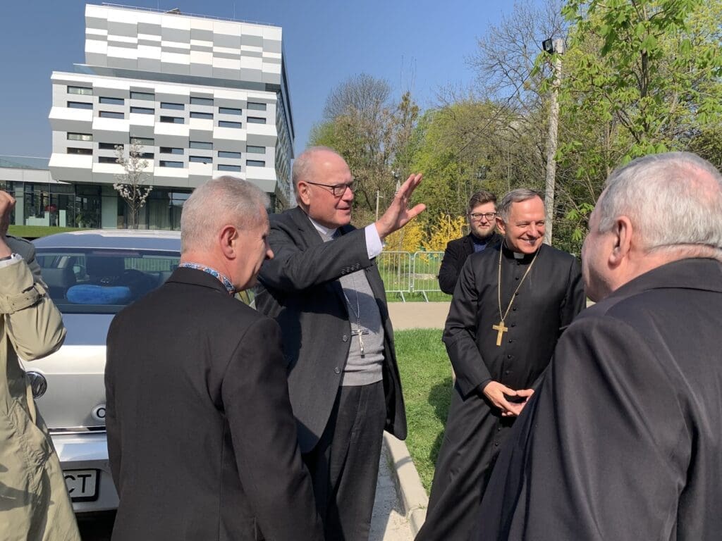 Cardinal Timothy Dolan, standing on a street, waves to other people.