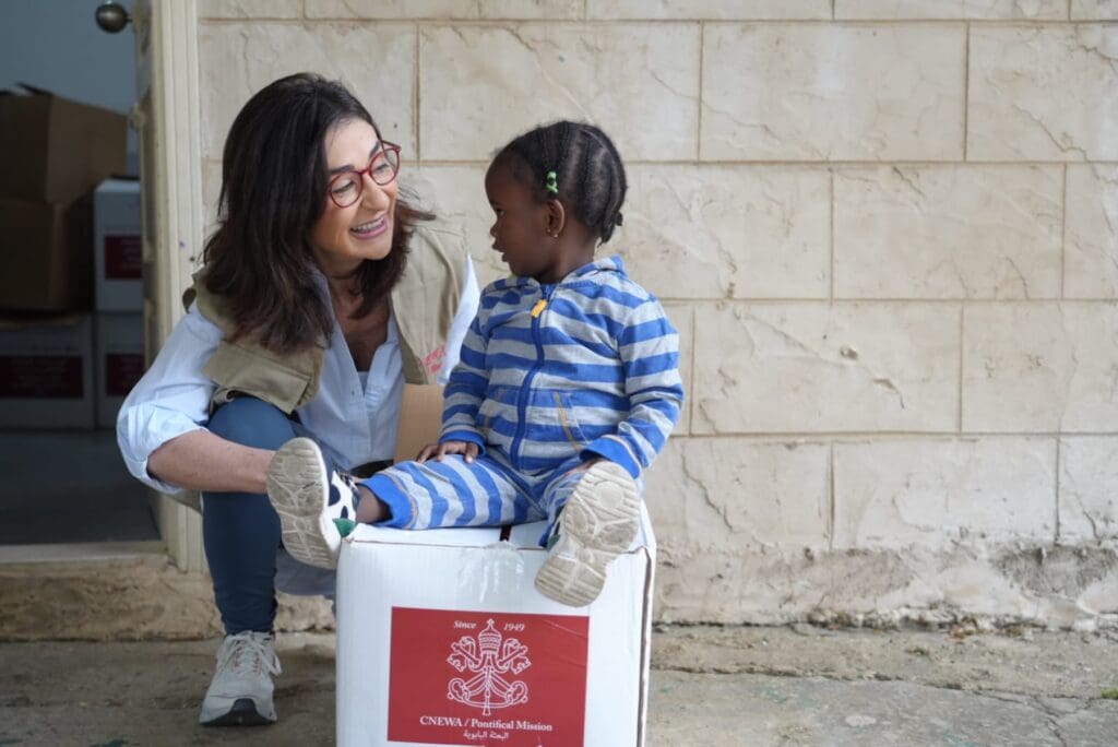 A woman crouching down smiles with a girl sitting on a box with a pontifical logo.