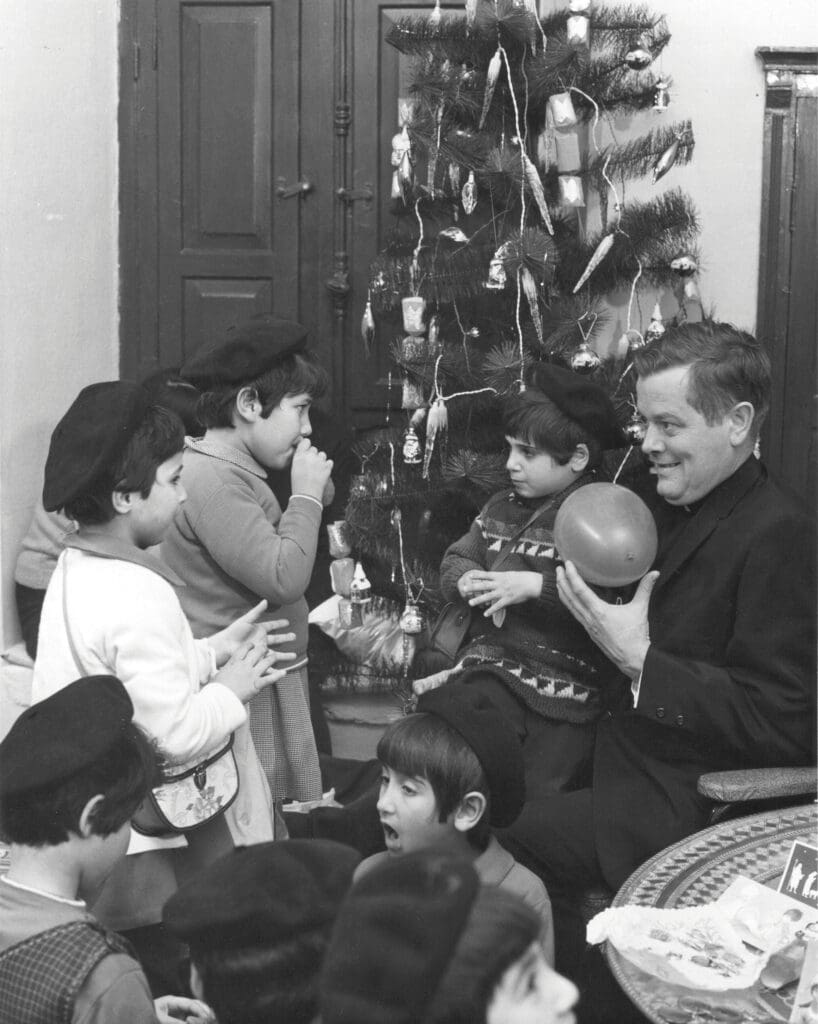 A priest plays with children next to a Christmas tree, on this black and white picture.