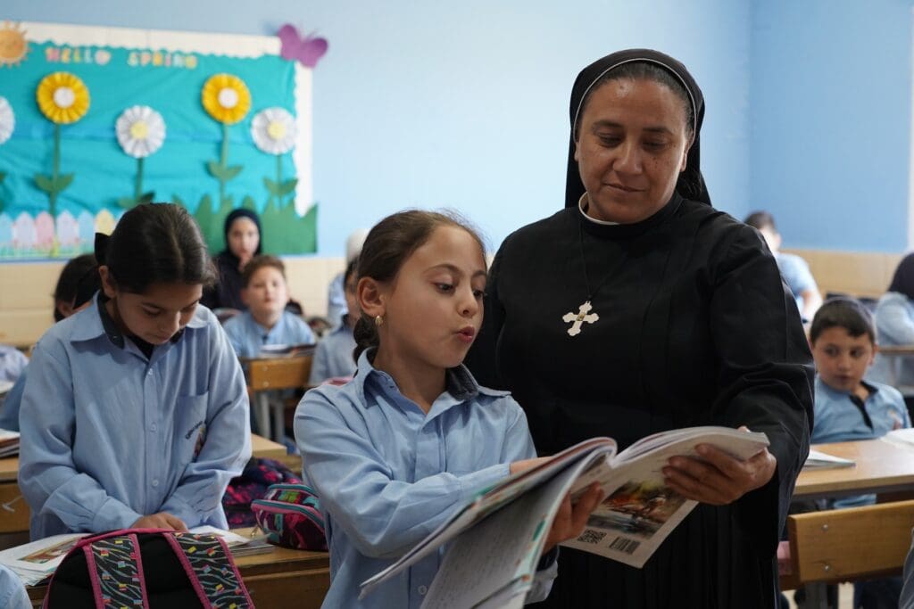 A religious teacher in a classroom next to a girl who is reading a book.